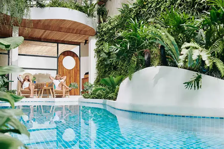 Modern luxury courtyard with a blue-tiled swimming pool, lush tropical plants on white curved walls, and two people relaxing in wooden chairs outside a contemporary home.